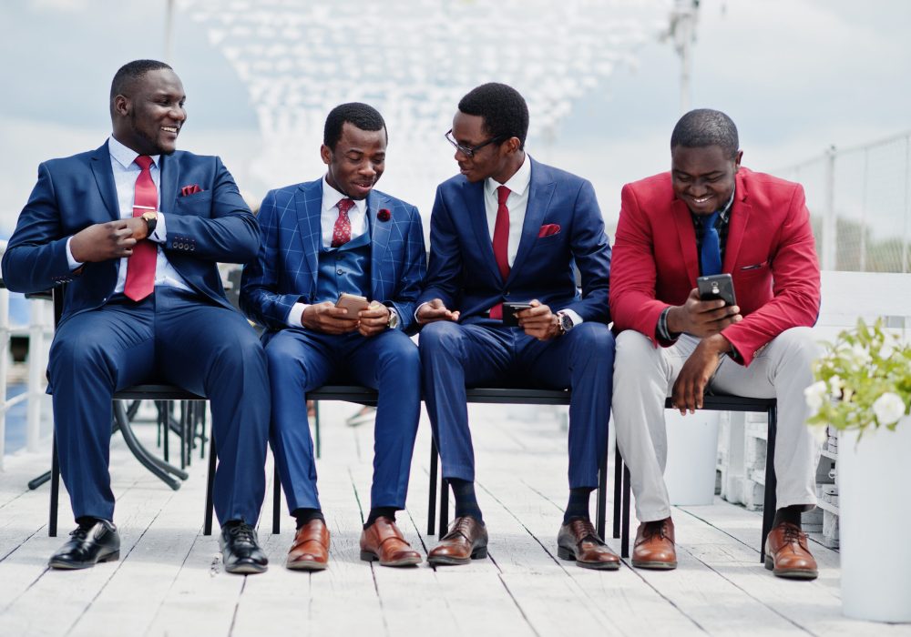 Group of four african american happy succesfull mans at suit. Rich black business mans sitting at chairs looking at mobile phones.